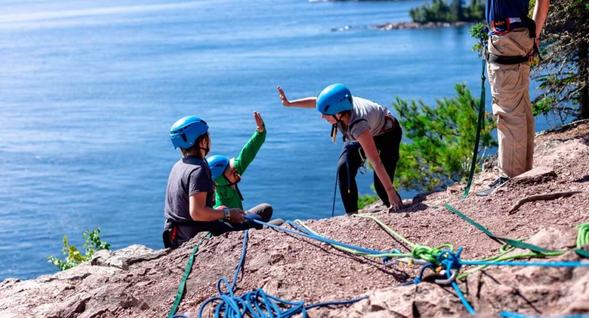 Two students wearing safety gear and secured by ropes high five on the edge of a cliff, high above a large blue lake. 